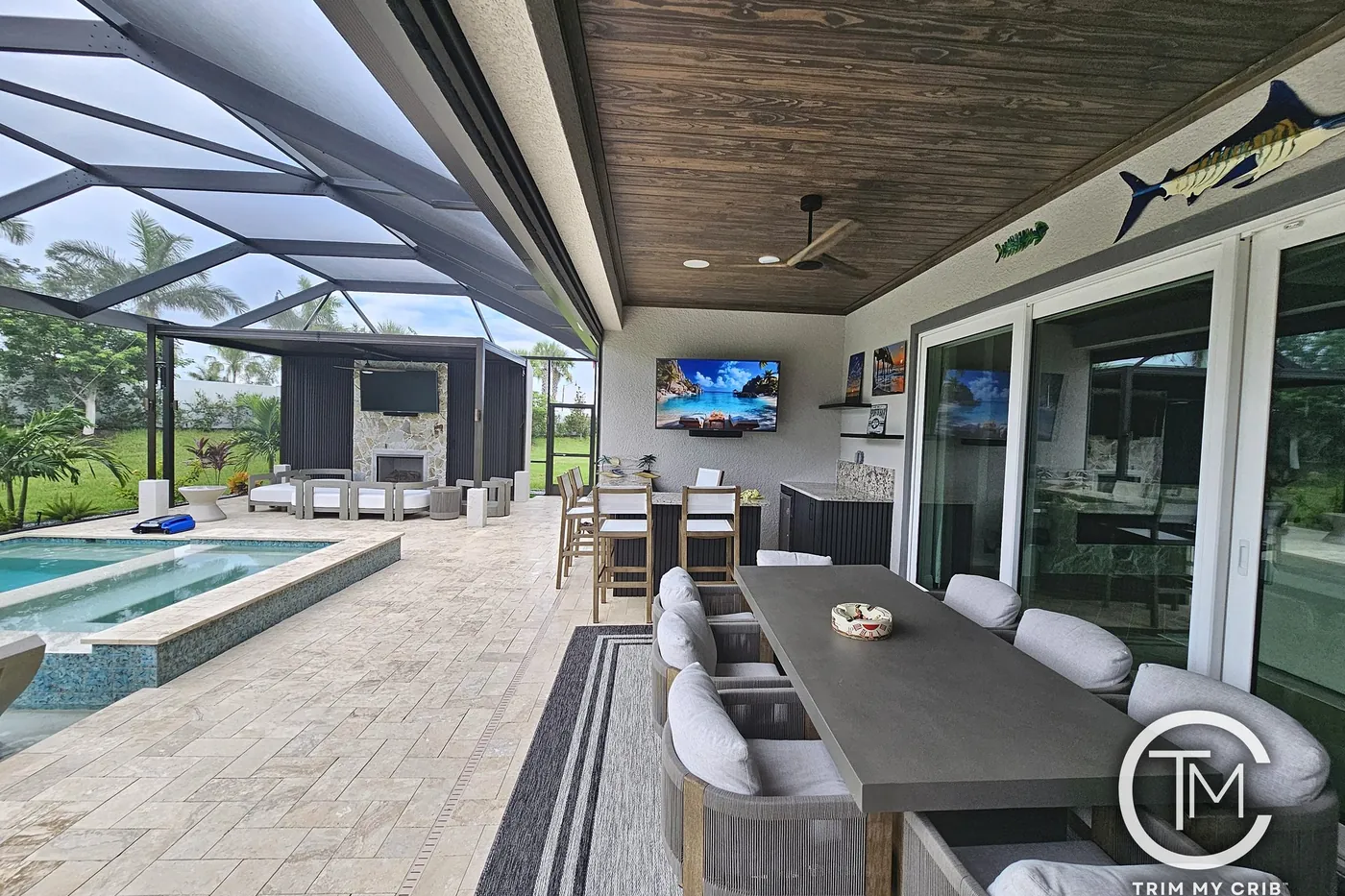 Outdoor kitchen and long dining table on a lanai under a stained wood ceiling beside the pool