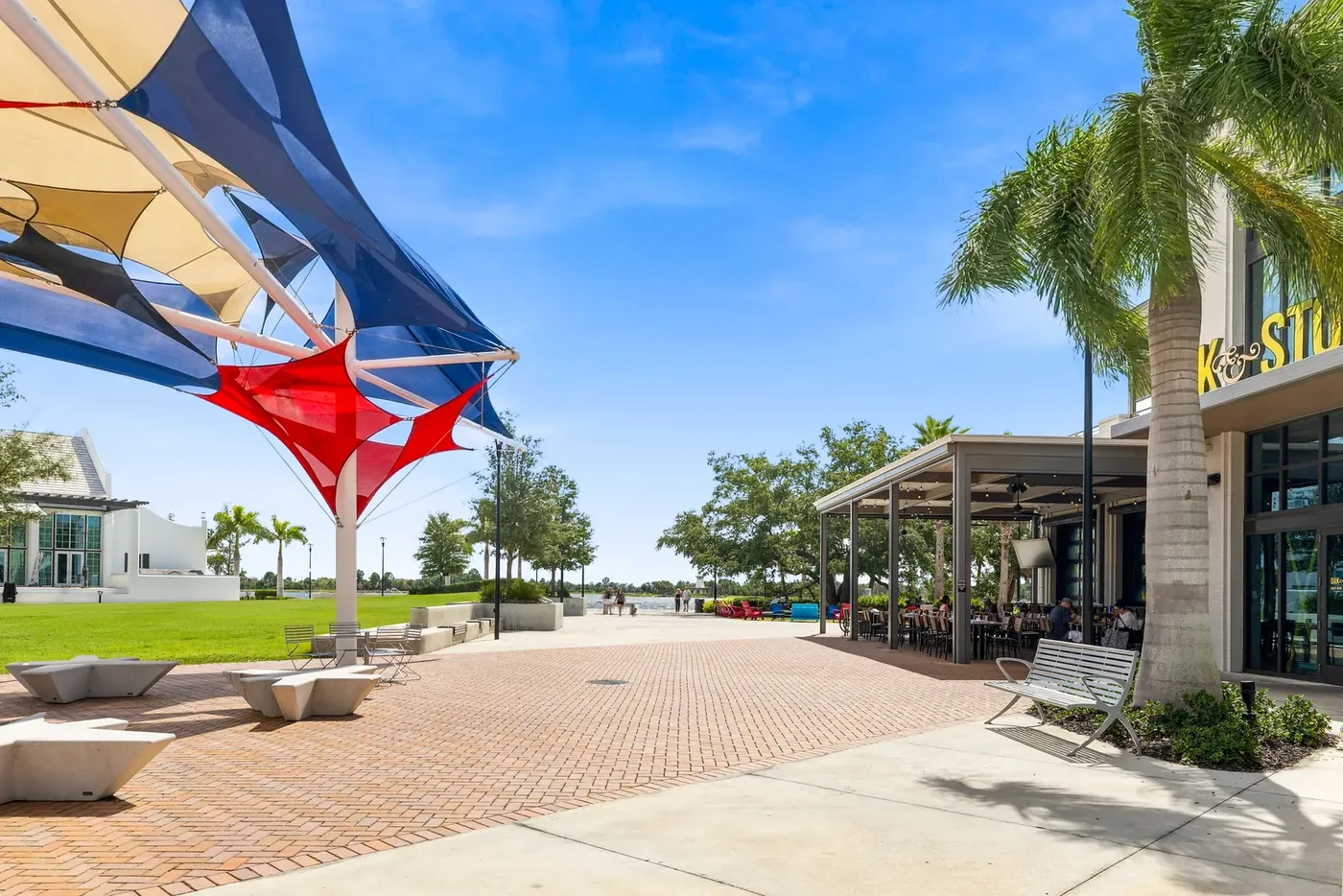 Red, white and blue shade sails over a brick plaza with a lawn and palms in Downtown Wellen