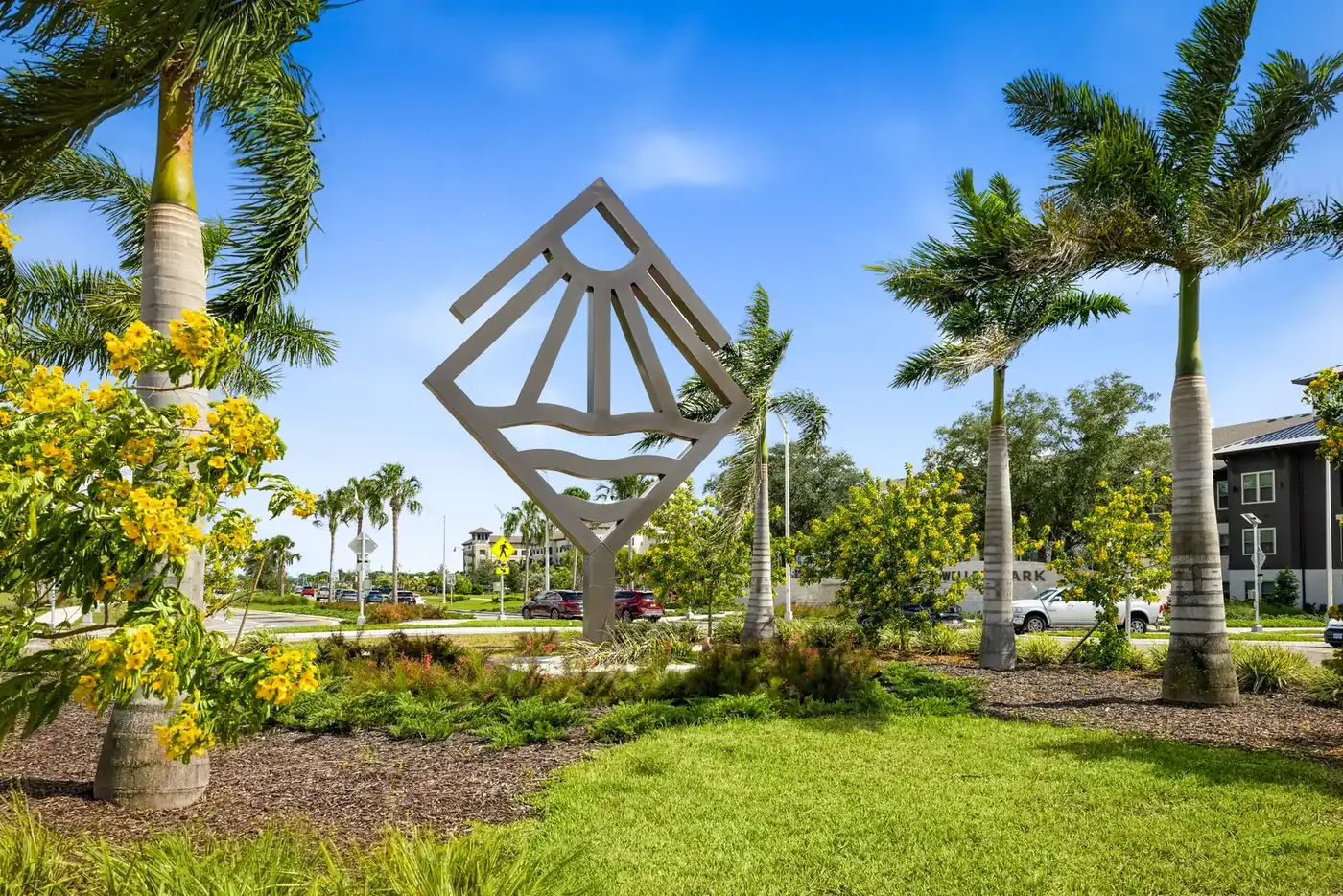 Geometric sun-and-waves sculpture in a landscaped garden with palms and yellow flowers in Wellen Park